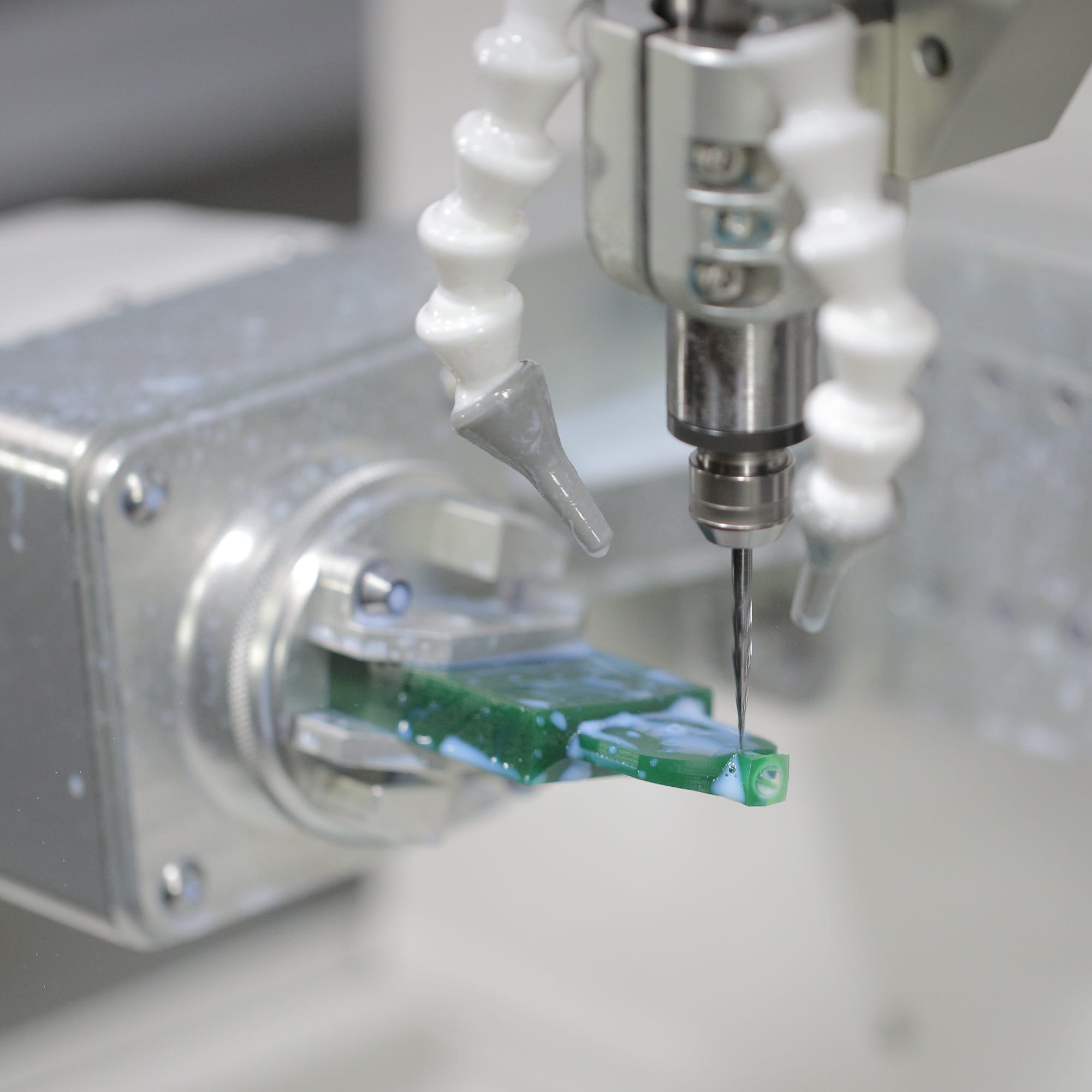 A slender micro end mill cutting a green workpiece held in a rotary fixture, with coolant nozzles aimed at the cut